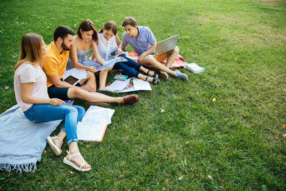 Group of cheerful students teenagers in casual outfits with note books and laptop are studying outdoors